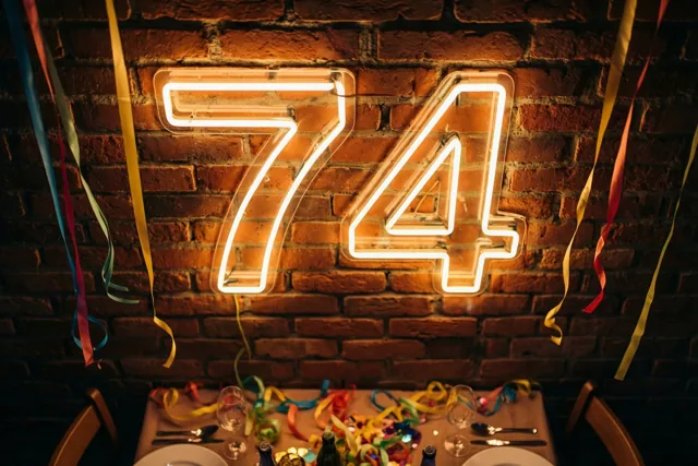 Een lichtgevend neongetal 74 hangt aan een bakstenen muur boven een feestelijk versierde tafel in warme oranje en gele tinten.