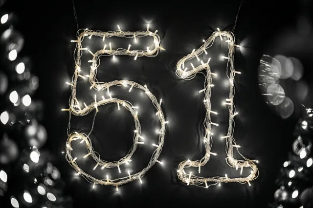 Warme lichtjes vormen het getal 51 tegen een donkere muur met onscherpe lichtjes op de achtergrond.
