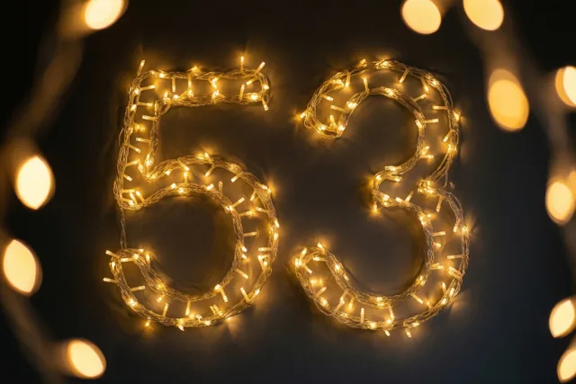 Warme lichtjes vormen het getal 53 op een donkere muur, met zachte lichtvlekken op de achtergrond.