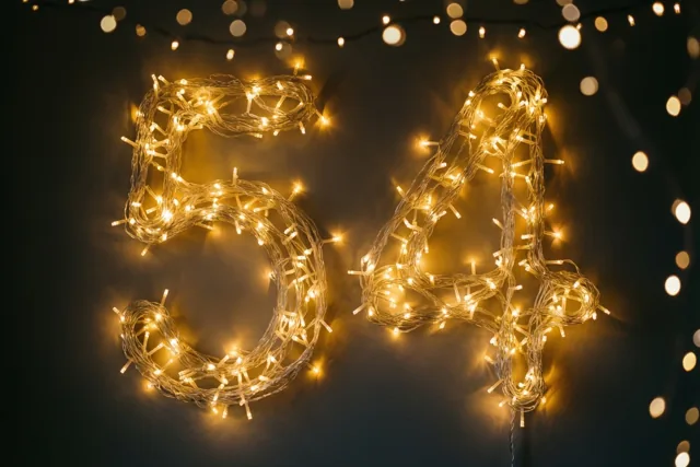 Warme lichtjes vormen het getal 54 tegen een donkere muur, met onscherpe lichtvlekken op de achtergrond.