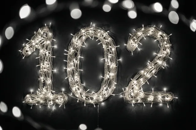 Warmwitte lichtslingers vormen het getal 102 tegen een donkere muur met onscherpe lichtjes op de achtergrond.