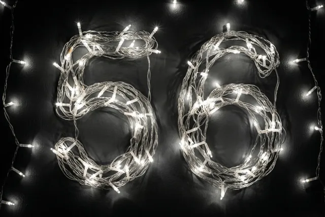 Warmwitte lichtjes vormen het getal 56 op een donkere muur, met onscherpe lichtvlekjes eromheen.
