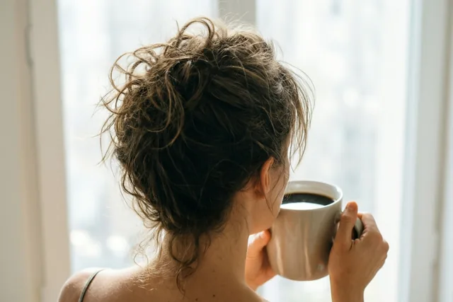 Een persoon met warrig haar is van achteren te zien met een grote koffiemok in de hand, kijkend door een raam in zacht daglicht.