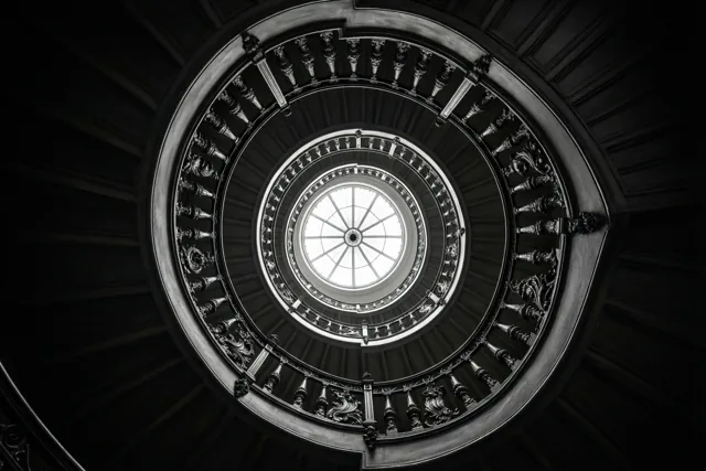 Een zwart-witfoto toont van onderaf een wenteltrap met donkere, sierlijke balustrades en treden rond een rond dakraam.