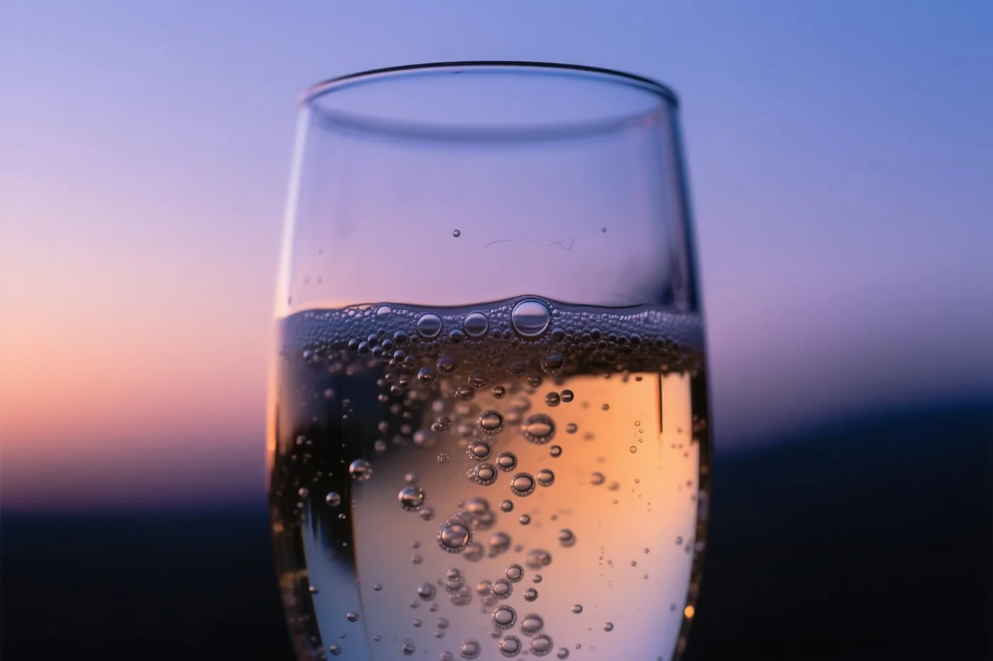 Een close-up toont een glas mousserende wijn met opstijgende belletjes voor een schemerhemel in paarse, oranje en gouden tinten.
