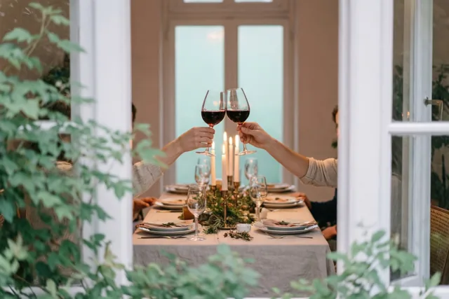Door een raam met een wit kader zijn twee handen te zien die met wijnglazen klinken boven een gedekte tafel met kaarsen en groen.
