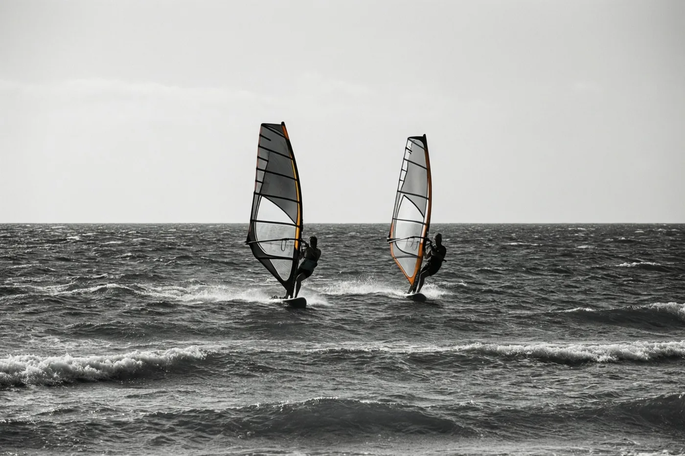 Twee windsurfers varen in de verte naast elkaar over een woelige zee op een zwart-witfoto.