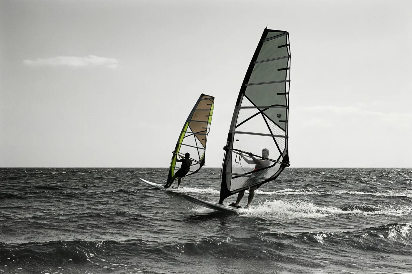 Twee windsurfers varen in de verte naast elkaar over een woelige zee op een zwart-witbeeld met een groen accent op één zeil.