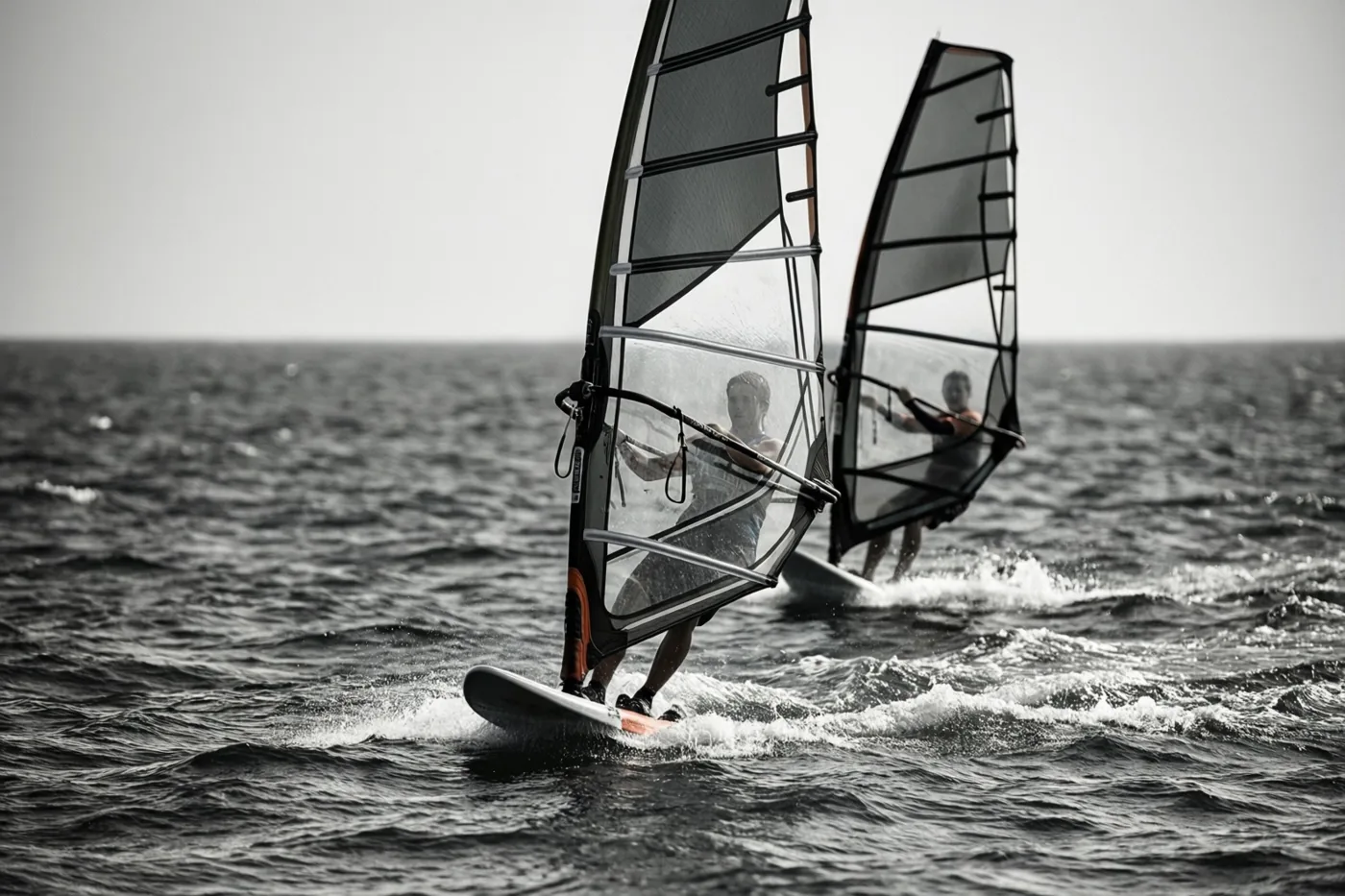 Twee windsurfers varen in de verte naast elkaar over een woelige zee, op een zwart-witbeeld met subtiele oranje accenten op de planken en zeilen.