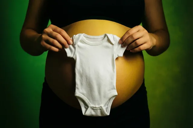Een zwangere vrouw houdt een klein wit babypakje voor haar buik tegen een groene achtergrond.