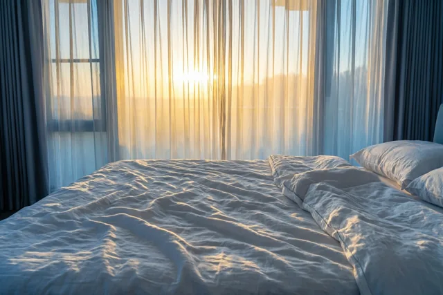 Gouden ochtendlicht valt door doorschijnende gordijnen op een bed met verkreukelde witte linnen lakens in een slaapkamer met witte, gouden en blauwe tinten.