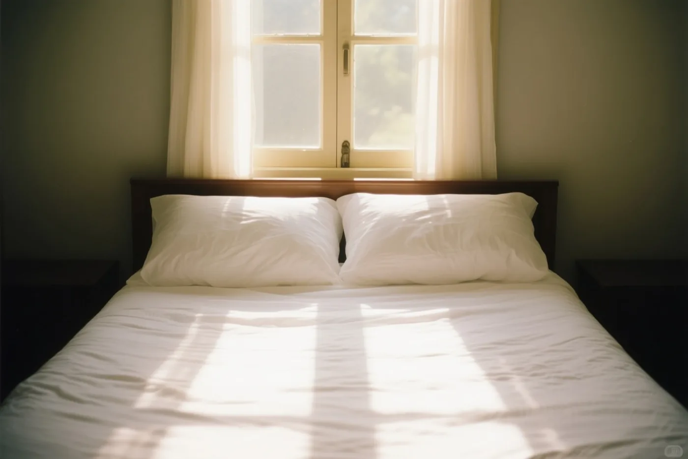 Een wit bed met twee kussens staat onder een raam met doorschijnende gordijnen in een kamer met grijsgroene muren en donker houten meubels, terwijl zonlicht over de sprei valt.
