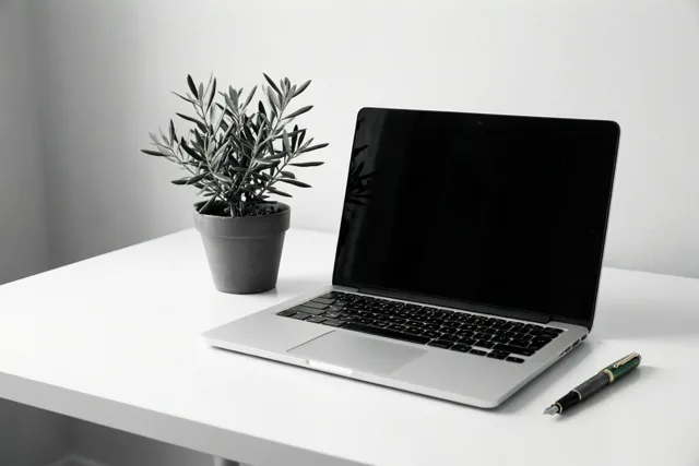 Op een opgeruimd wit bureau staan een zilverkleurige laptop met zwart scherm en een klein olijfplantje in een pot, naast een groen-zwarte vulpen.