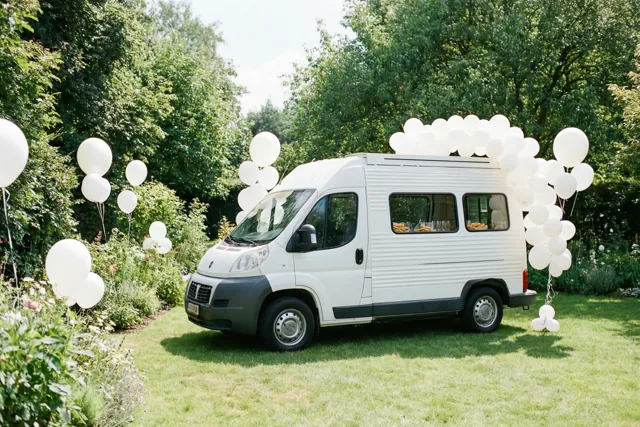 Een wit busje ingericht als frietkot staat op het gras in een tuin, omringd door trossen witte ballonnen en groene bomen in helder daglicht.