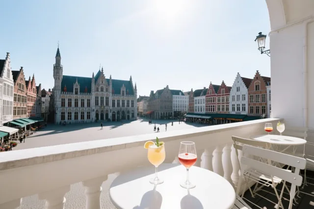 Een wit terras met kleurrijke drankjes op de tafels kijkt uit op een zonnig Vlaams plein met historische gebouwen en een centraal stadhuis onder een blauwe hemel.