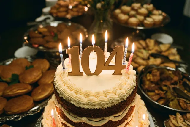 Een stapeltaart met wit glazuur, brandende kaarsen en een papieren 104 staat op een buffet tussen schalen met gebak en desserts.