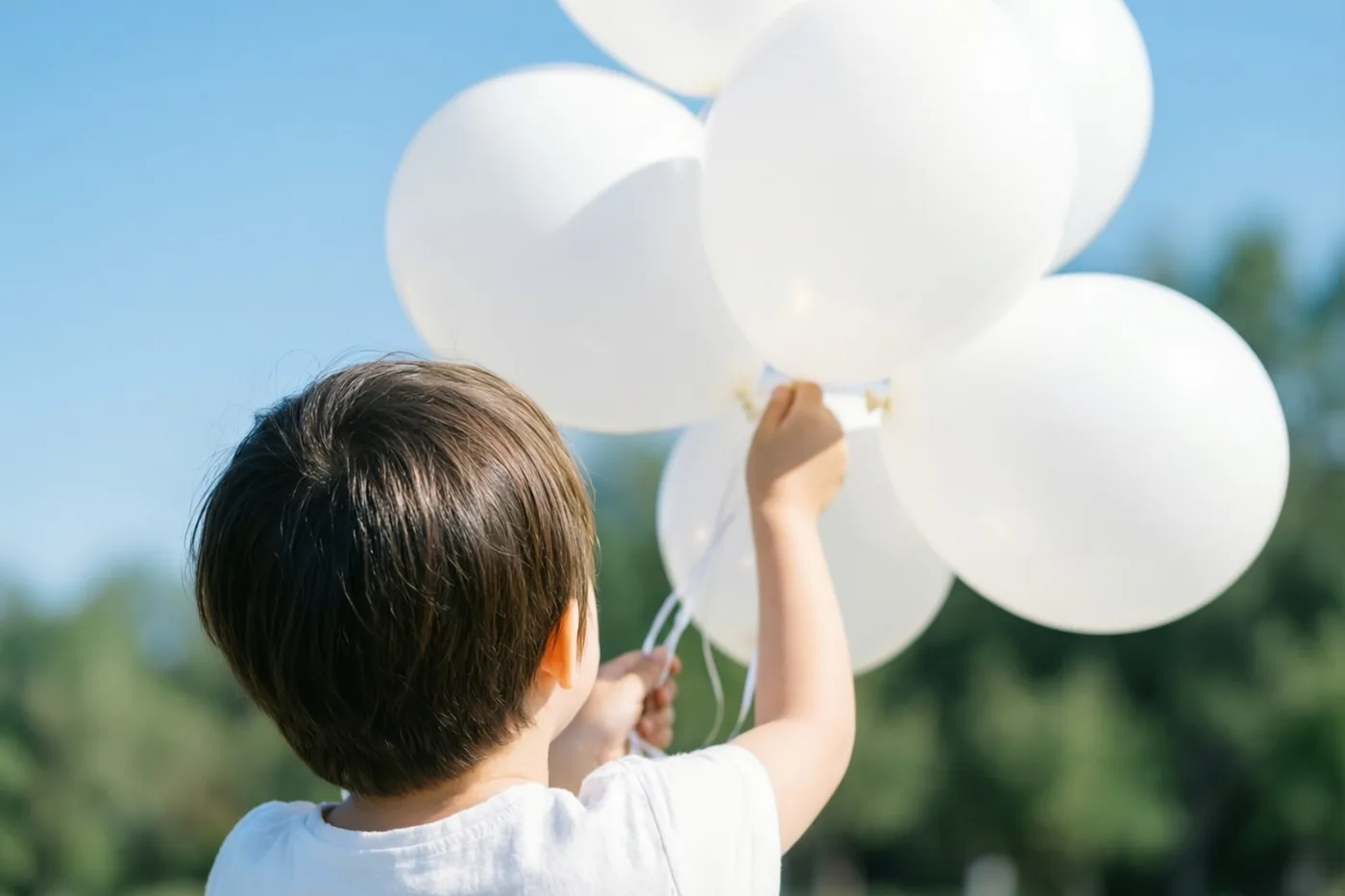 Een jong kind houdt buiten met de rug naar de kijker een tros witte ballonnen vast tegen een blauwe hemel en onscherp groen.