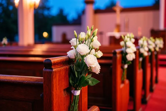 Witte bloemen sieren de uiteinden van houten kerkbanken in warm gouden licht, met een blauwe schemerlucht zichtbaar door de ramen.