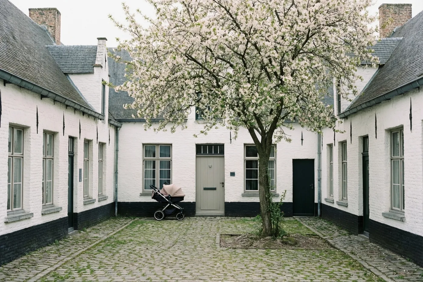 Een kinderwagen staat bij een deur op een binnenplein met kasseien, witte begijnhofhuisjes met donkere leien daken en een boom met witte bloesems.
