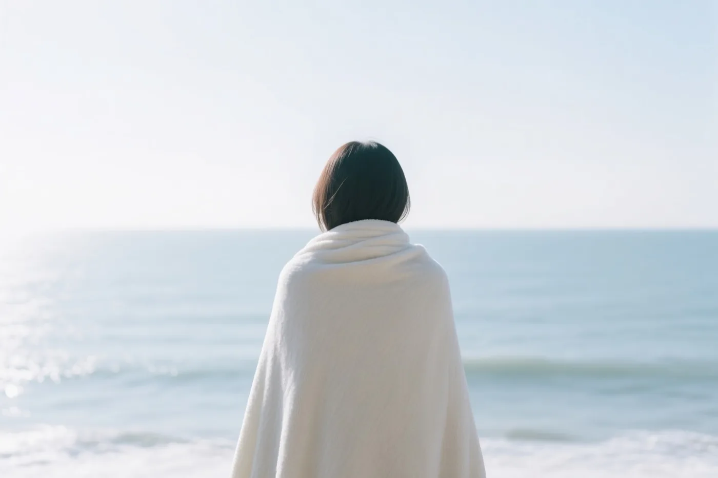 Een persoon met kort donker haar staat met de rug naar de kijker in een witte deken op het strand en kijkt naar een kalme zee onder een heldere hemel.