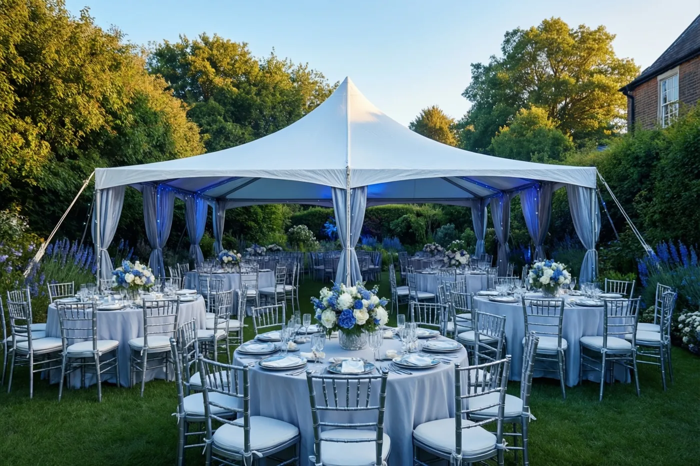 Een witte tent in een groene tuin heeft blauwe verlichting langs de randen, ronde tafels met witte kleden en bloemstukken, en zilverkleurige stoelen.
