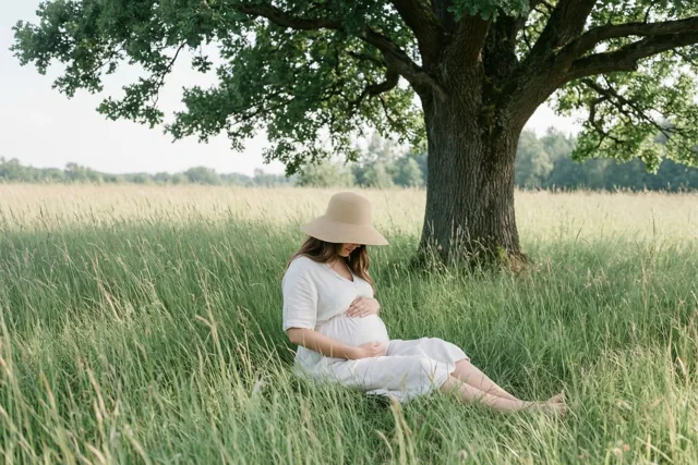 Een zwangere vrouw in een witte jurk en een strohoed zit in hoog groen gras onder een grote eik, met haar handen op haar buik in zacht licht.