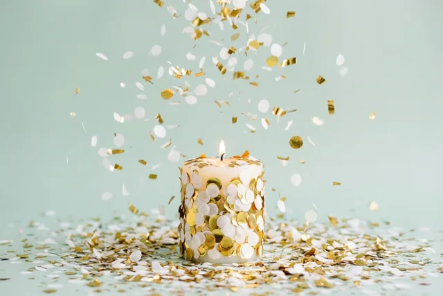 Een brandende witte kaars met witte en gouden confetti staat op een lichtgroen oppervlak, met vallende confetti tegen een lichtgroene achtergrond.