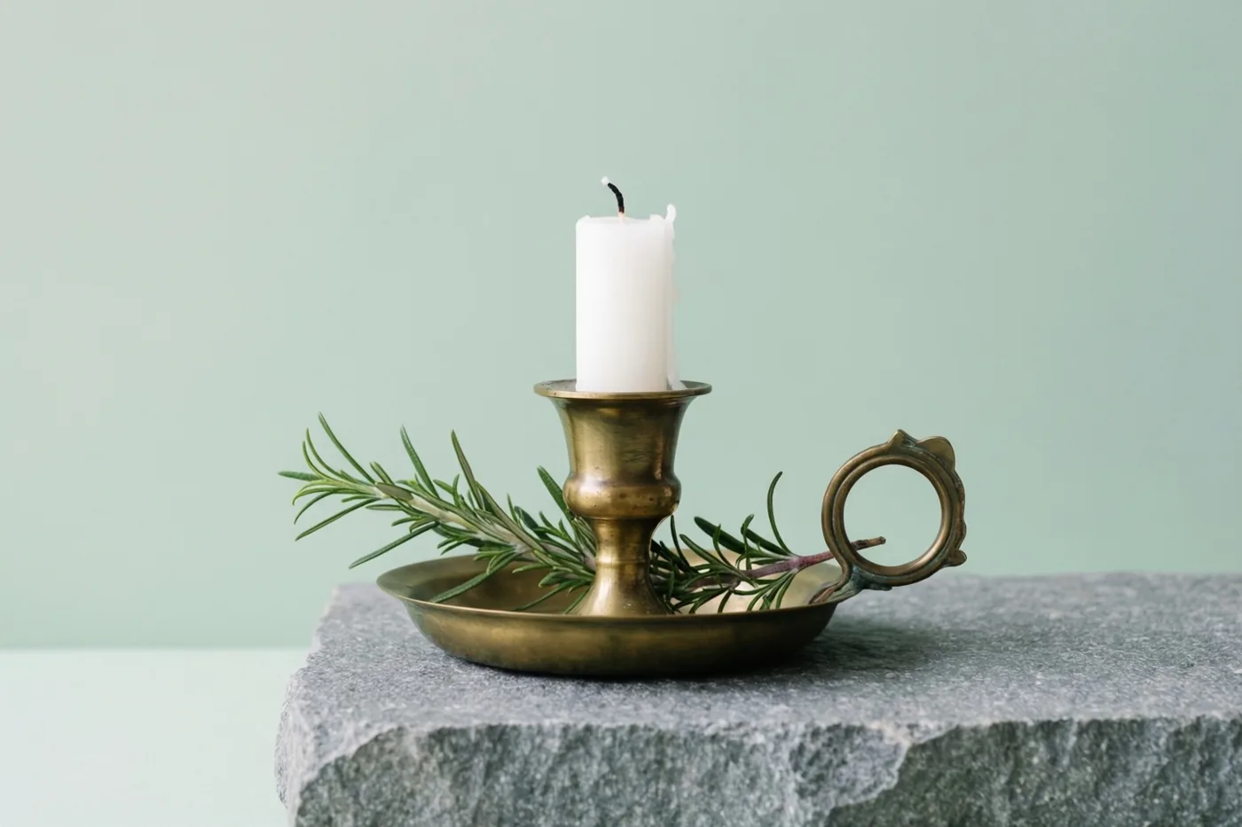 Een zware messing kandelaar met een dikke witte kaars en een takje rozemarijn staat op een grijs stenen oppervlak tegen een effen lichtgroene achtergrond.