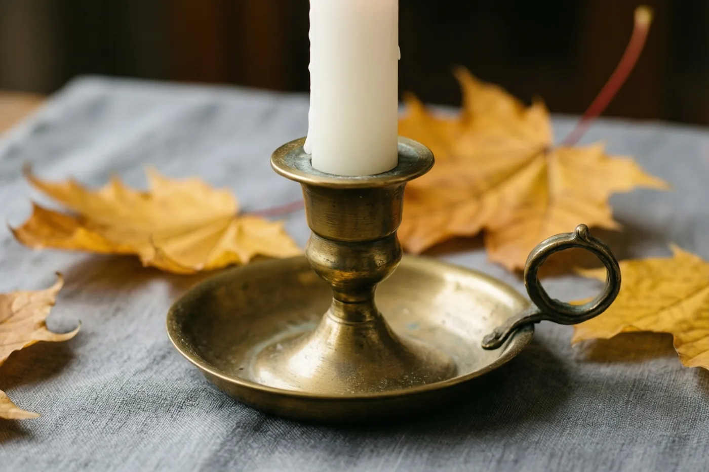 Een hoge witte kaars staat in een messing kandelaar op grijs linnen, met gouden herfstbladeren eromheen.