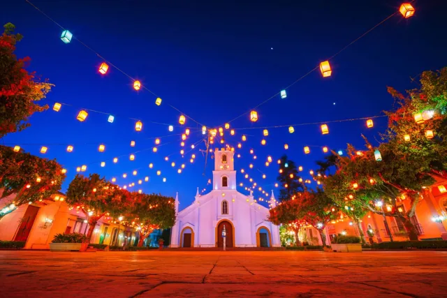 Een witte kerk staat 's nachts op een plein met slingers brandende lantaarns, in oranje en rood licht onder een diepblauwe lucht.