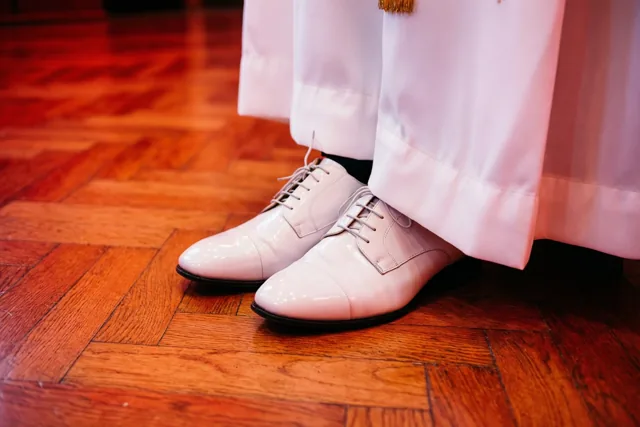 Een paar witte lakschoenen staat naast een wit communiegewaad op een glanzende parketvloer in warm licht.