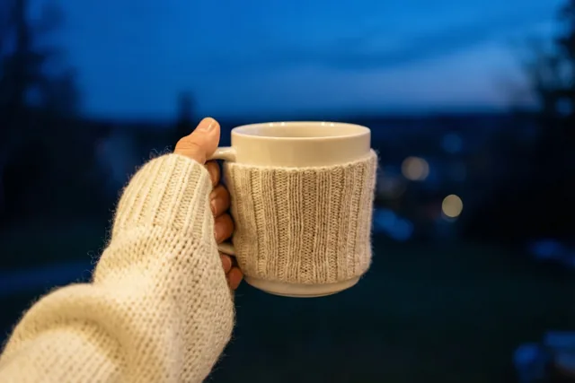 Een hand uit een crèmekleurige gebreide mouw houdt een witte mok met een bijpassende gebreide hoes vast tegen een onscherpe blauwe avondlucht.