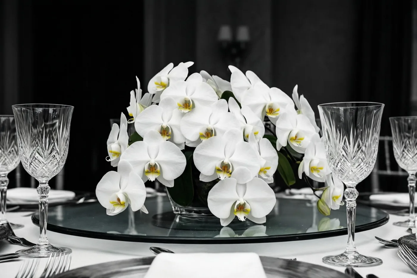 Witte vlinderorchideeën staan op een glazen feesttafel tussen kristallen wijnglazen tegen een donkere achtergrond.