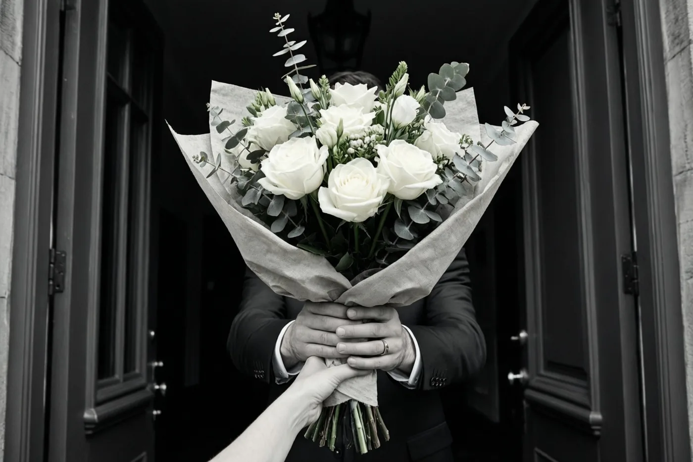 Iemand in een donker pak reikt aan een open voordeur een boeket witte rozen en eucalyptus in linnenpapier aan, terwijl een andere hand het aanneemt, op een zwart-witfoto.