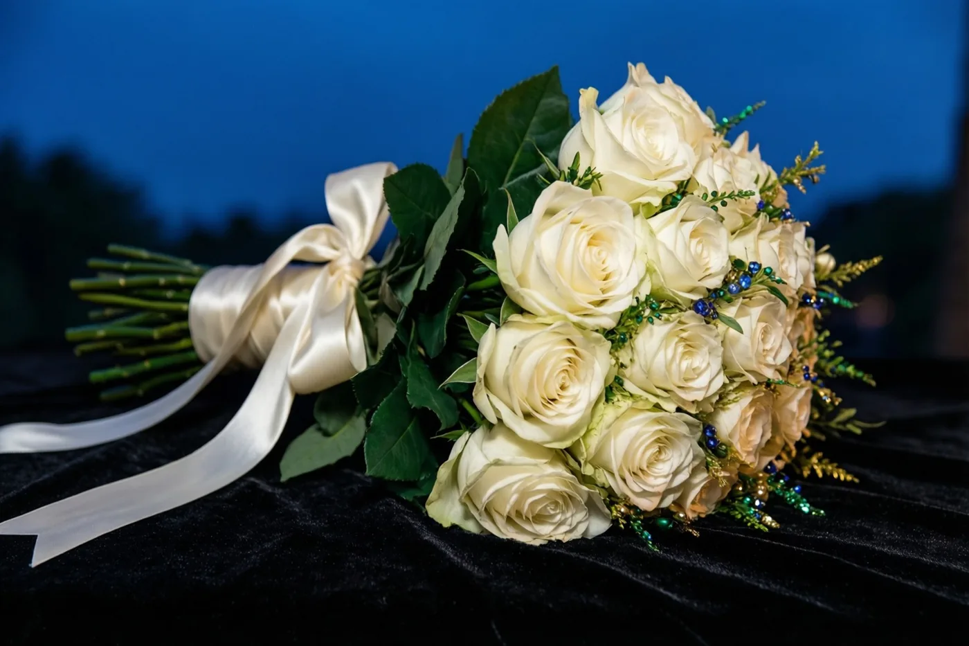 Een boeket witte rozen met een ivoorkleurig satijnen lint ligt op zwart fluweel tegen een diepblauwe schemerlucht.