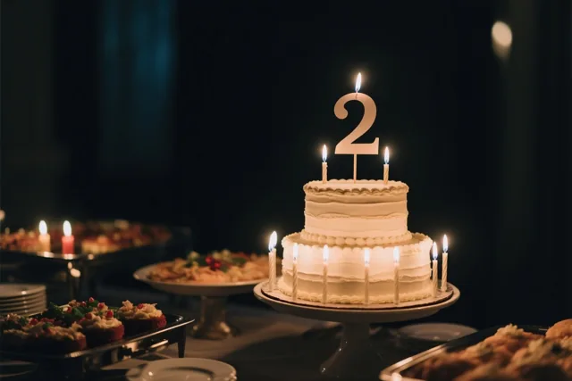 Een witte taart met twee verdiepingen, een groot papieren cijfer 2 en brandende kaarsjes staat op een buffet, met desserts en kaarsen in een schemerige kamer.