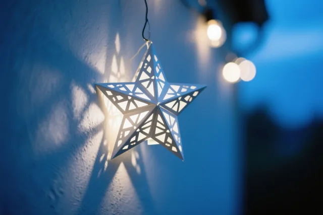 Een hangende witte sterlantaarn met fijne uitsparingen werpt een lichtpatroon op een blauwe muur met structuur, met warme lichtjes op de achtergrond.
