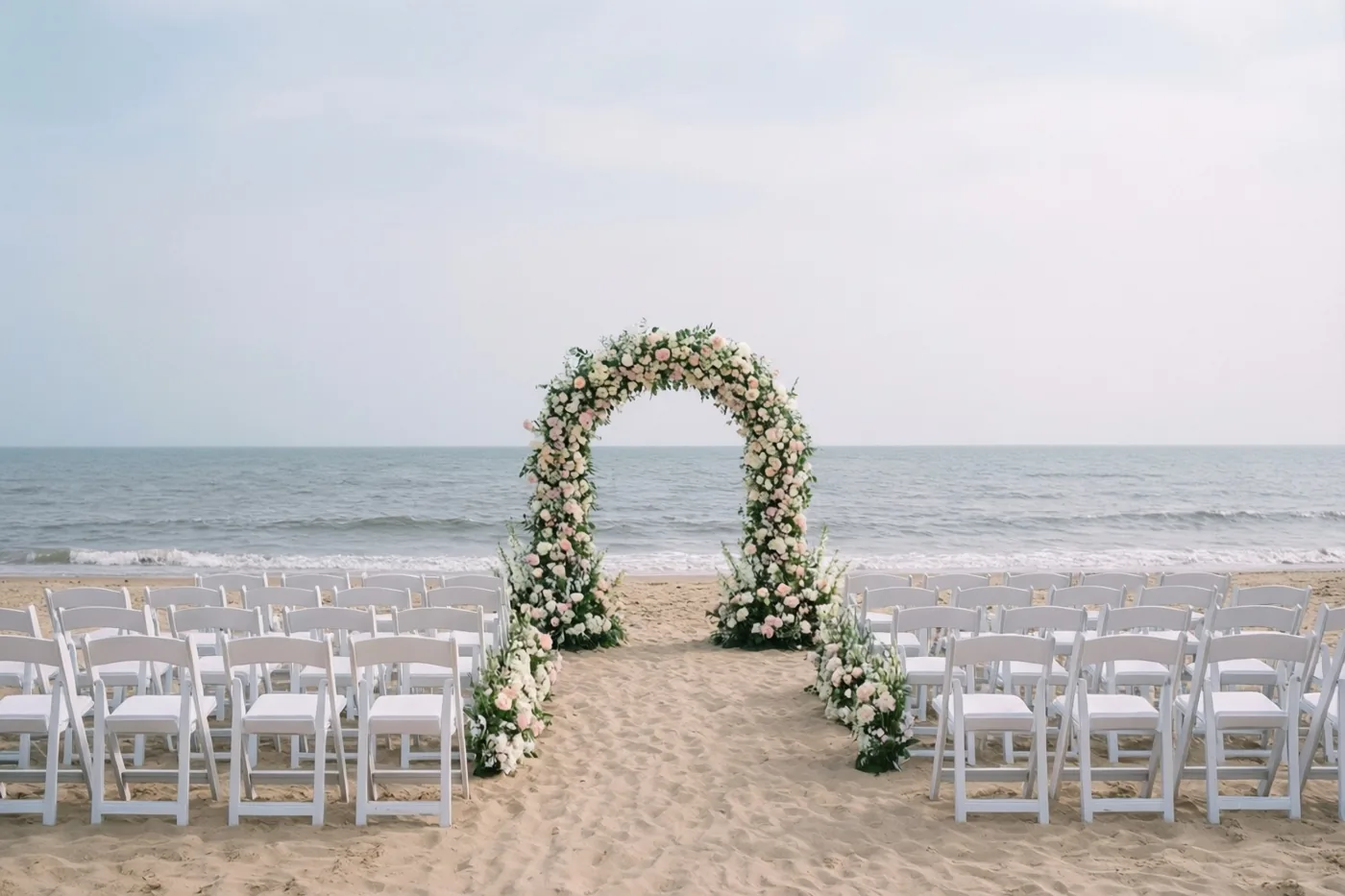 Rijen witte stoelen staan op een zandstrand tegenover een boog met roze en witte bloemen, met de grijsblauwe zee onder een bleke hemel erachter.