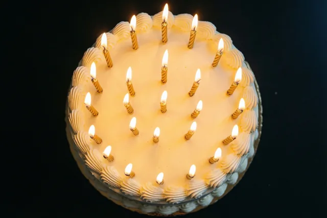 Van bovenaf is een ronde taart met wit glazuur en een kring brandende gouden kaarsen te zien tegen een zwarte achtergrond.