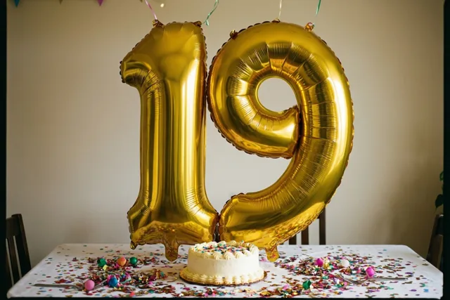 Gouden cijferballonnen vormen 19 boven een tafel met een tafelkleed, kleurrijke confetti en een kleine witte taart, tegen een effen muur.