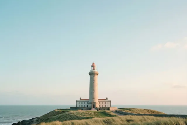 Een hoge witte vuurtoren staat op een met gras begroeide kustklif boven zee onder een heldere hemel in zacht, warm licht.