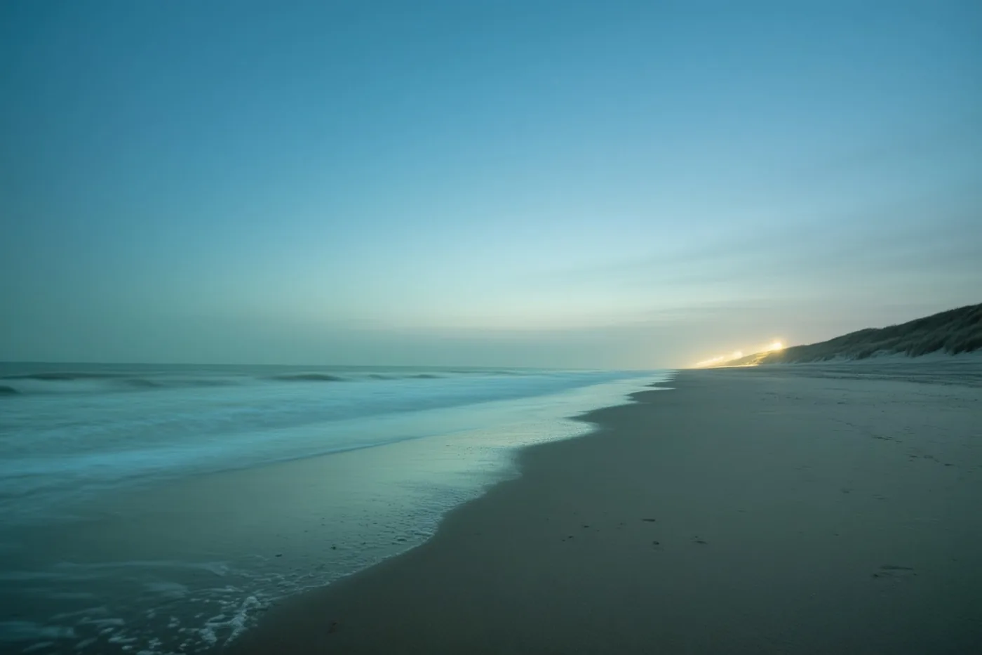 Zacht vervaagde golven rollen op een verlaten strand onder een blauwe lucht, met duinen rechts en een lichte gloed aan de verre horizon.