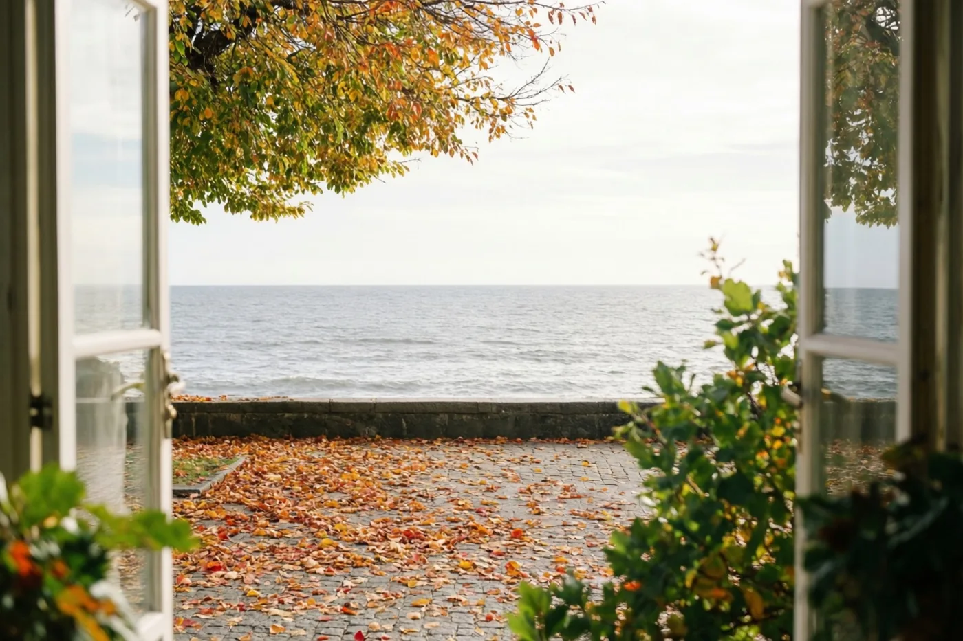 Door een open raam is een promenade met herfstbladeren langs een grijze zee te zien, met bruin, oranje en groen gebladerte in beeld.