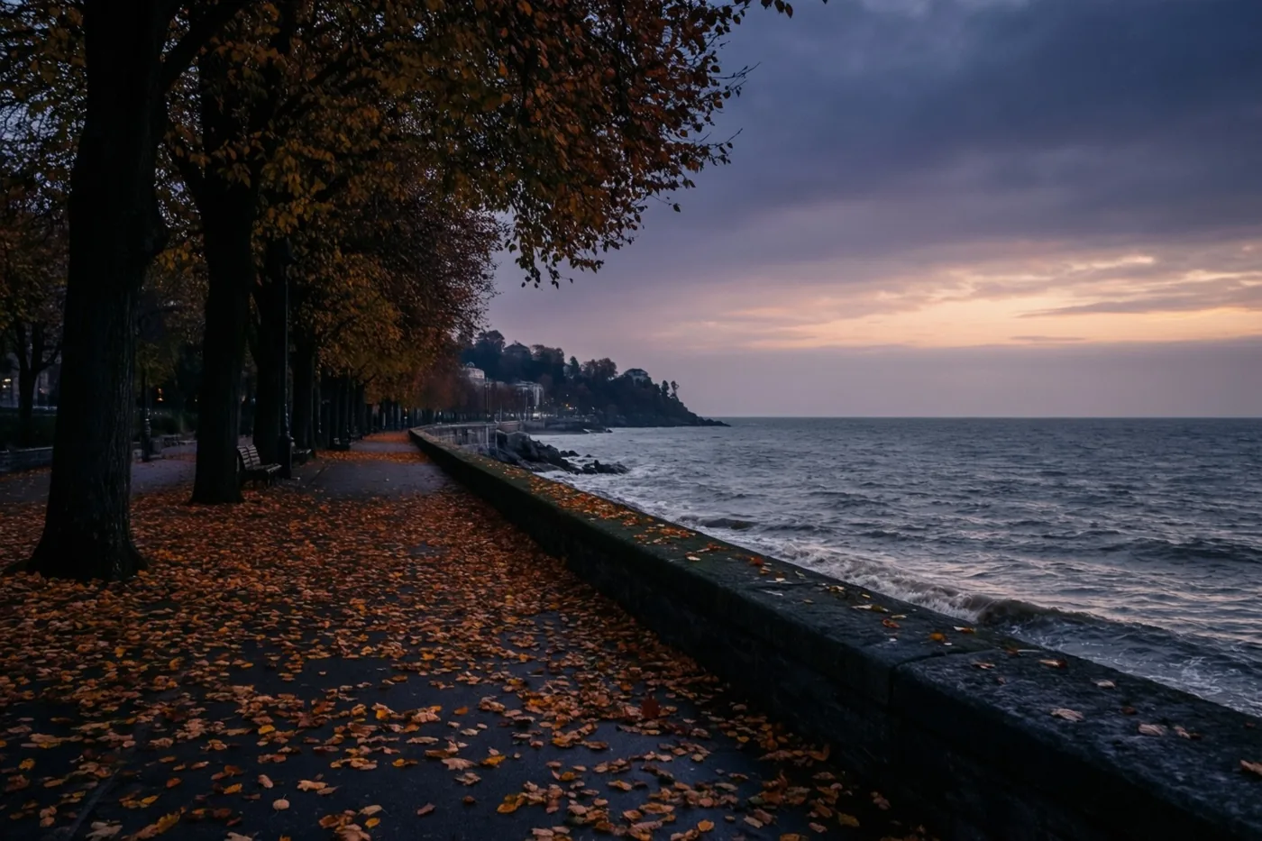 Een promenade vol gevallen herfstbladeren loopt langs een grijze zee onder een bewolkte schemerhemel, met links bomen met oranje en geel blad.