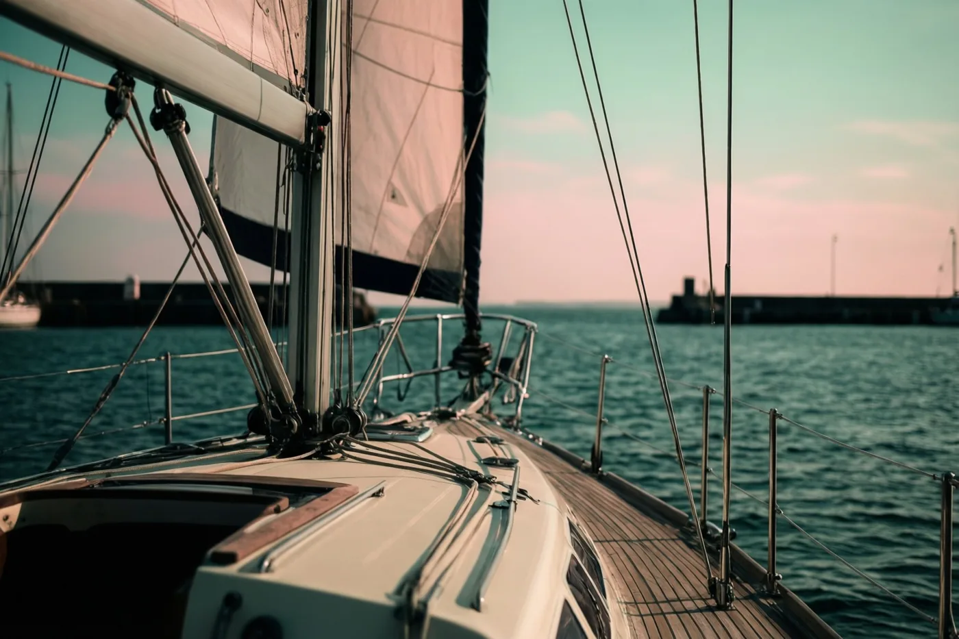 Vanaf het dek van een zeilboot met crèmekleurige oppervlakken is te zien hoe die de haven verlaat over blauwgroen water onder een bleke hemel.