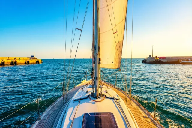 Een zeilboot met een wit zeil vaart tussen stenen golfbrekers de haven uit en laat een kielzog achter op het blauwe water in gouden zonlicht.