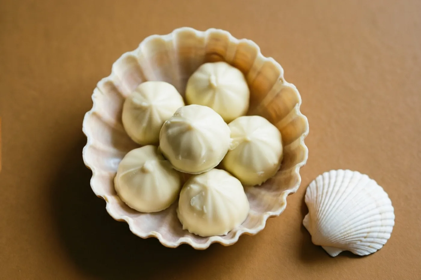 Zes witte chocoladepralines liggen in een schelpvormig schaaltje met geschulpte rand op een bruine ondergrond, naast een kleine witte doopschelp.