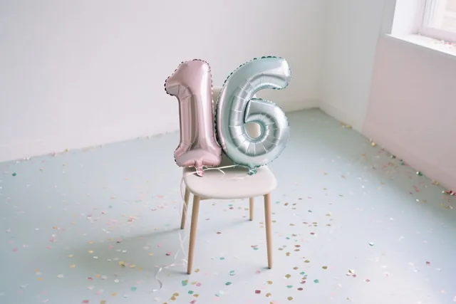 Twee folieballonnen vormen 16 aan een witte stoel in een sober ingerichte kamer met lichtblauwe muren en vloer, met confetti op de grond.