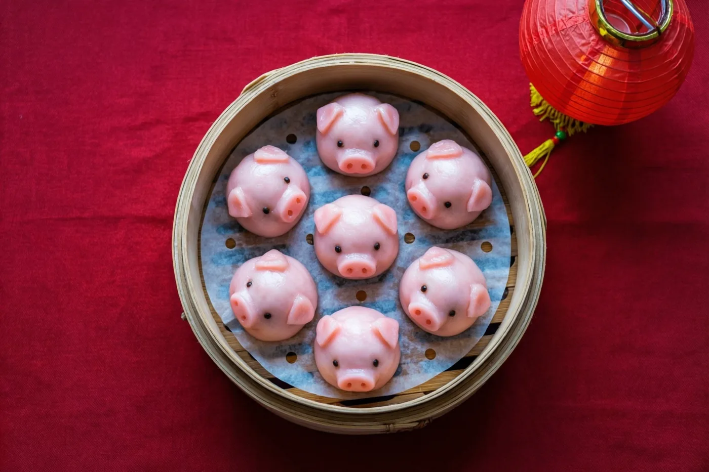 Zeven roze dumplings in de vorm van varkentjes liggen in een bamboe stoommand op een rood tafelkleed, met een rood lantaarntje ernaast.