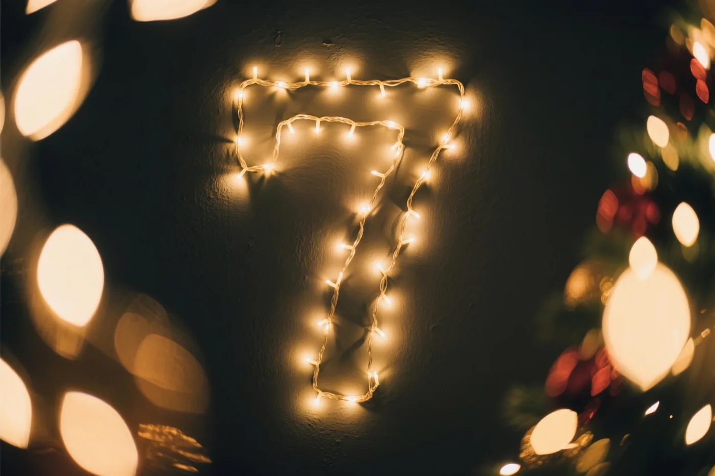 Warme lichtjes vormen het cijfer 7 op een donkere muur, omringd door onscherpe feestlichtjes.